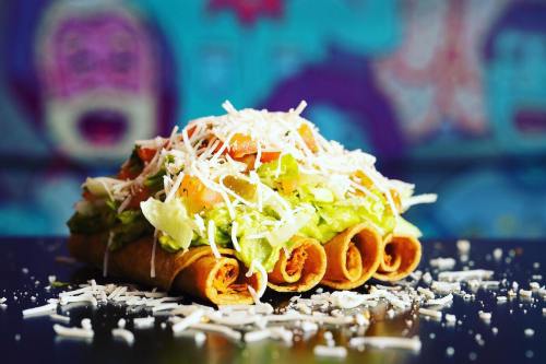 A colorful plate of rolled tacos topped with lettuce, tomatoes, guacamole, and shredded cheese, against a vibrant background.