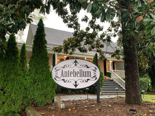A charming house with a sign reading "Antebellum," surrounded by greenery and trees on a cloudy day.