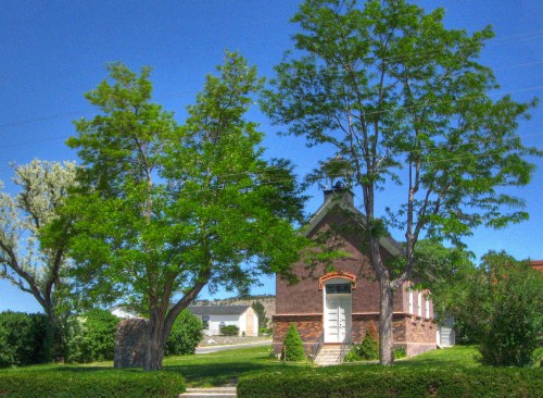 A brick building surrounded by lush green trees and shrubs under a clear blue sky.