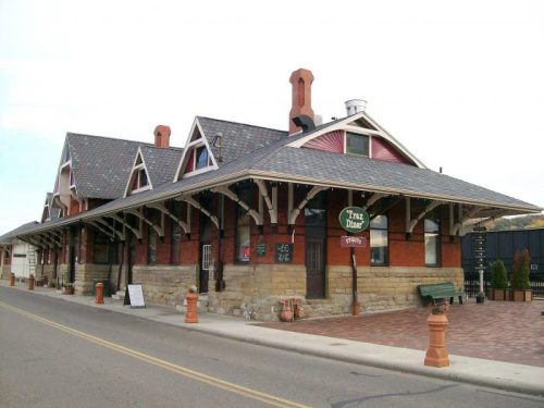 Historic train station with a brick facade, peaked roofs, and a welcoming sign, set along a quiet street.