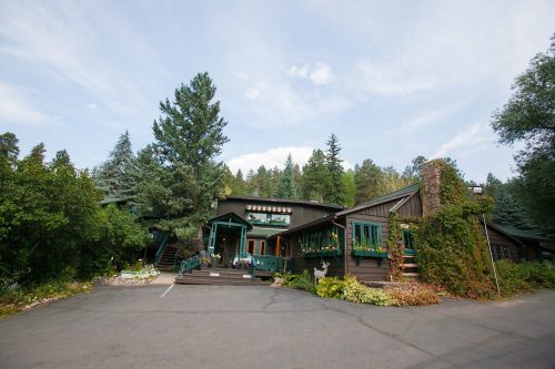 A rustic wooden lodge surrounded by trees and greenery, with a welcoming entrance and a clear blue sky above.
