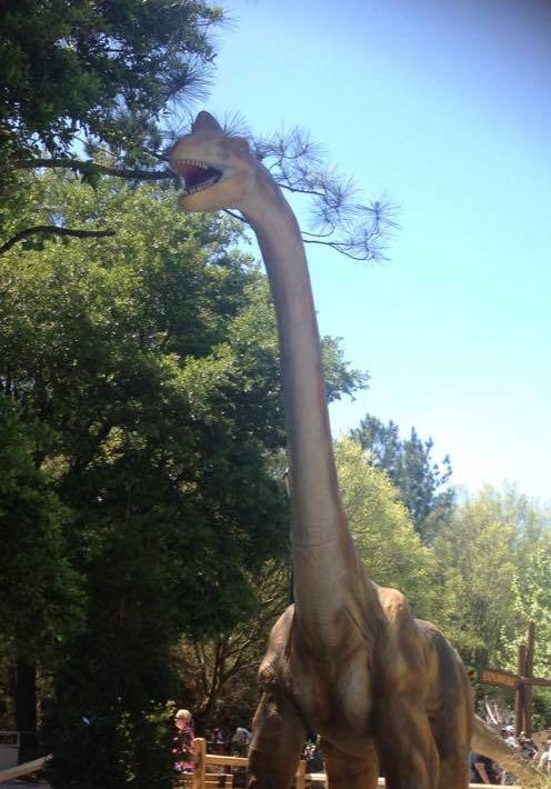 A child stands next to a tall dinosaur statue in a park, surrounded by trees and other visitors.