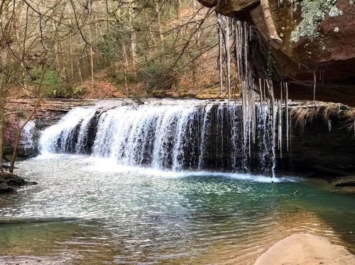 A serene waterfall cascades over rocks into a clear pool, surrounded by lush greenery and icicles hanging from above.