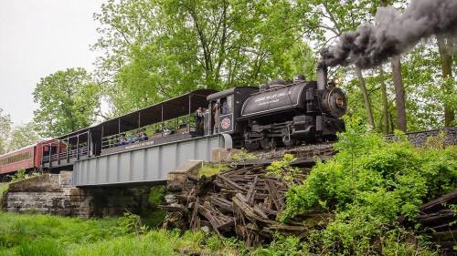 A vintage steam train crosses a bridge surrounded by lush greenery, with passengers visible in an open car.