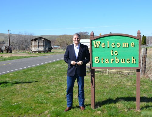 A man stands beside a colorful "Welcome to Starbuck" sign, with a rural landscape and an old building in the background.