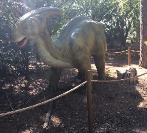 A child stands near a life-sized dinosaur statue in a wooded area, with a sign visible in the background.