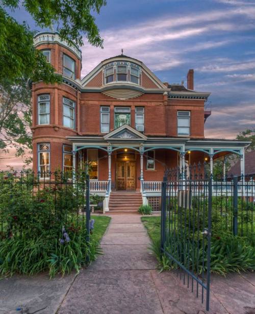 A large, historic red brick house with a porch, surrounded by greenery and a wrought iron gate, under a colorful sky.