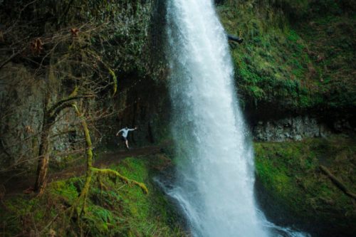 A person joyfully poses near a cascading waterfall surrounded by lush greenery and rocky terrain.
