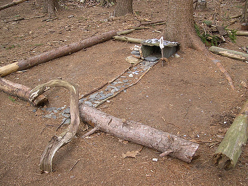 A small, rustic structure made of stones and wood, set in a forest clearing with fallen branches around it.