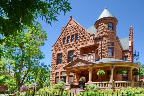 A large, ornate red stone house with a turret, surrounded by greenery and flowers under a clear blue sky.