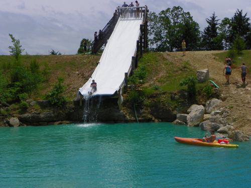 A water slide descends into a turquoise pond, with people enjoying the slide and kayaking nearby.
