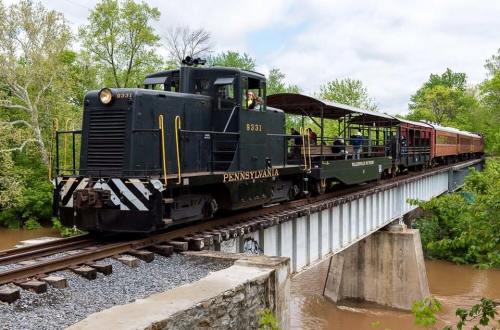 A vintage train crosses a bridge surrounded by greenery, with a mix of passenger and freight cars.