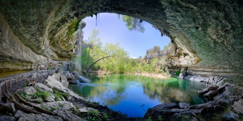 A scenic view of a cave opening overlooking a tranquil green lake surrounded by lush trees and rocky terrain.