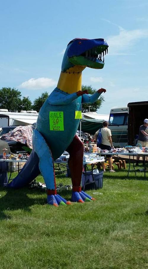 A colorful dinosaur sculpture stands in a field, with a "Sale" sign on it, surrounded by tables of items for sale.