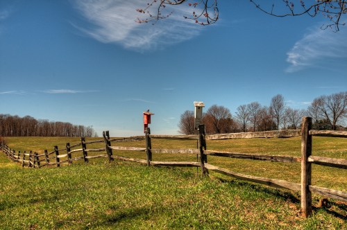 A scenic landscape featuring a wooden fence, birdhouses, and a clear blue sky with scattered clouds.