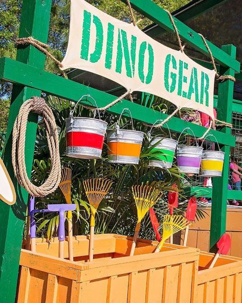 Colorful buckets and tools hang under a sign reading "DINO GEAR," set against a vibrant outdoor backdrop.