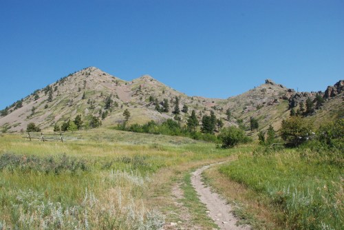 A dirt path winds through a grassy landscape, leading to rocky hills under a clear blue sky.