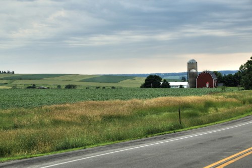 A rural landscape featuring green fields, a red barn, and a silos under a cloudy sky along a winding road.