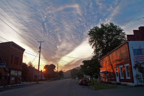 A quiet street at sunrise, with buildings on either side and dramatic clouds in the sky.