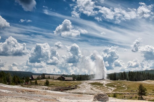 A geyser erupts in a scenic landscape with fluffy clouds and a distant building surrounded by greenery.