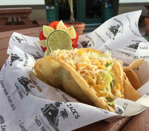 A basket of three tacos filled with cheese and lettuce, garnished with lime and tomato slices, on branded paper.