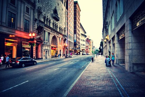 A quiet city street lined with buildings, shops, and a few pedestrians, illuminated by warm evening light.