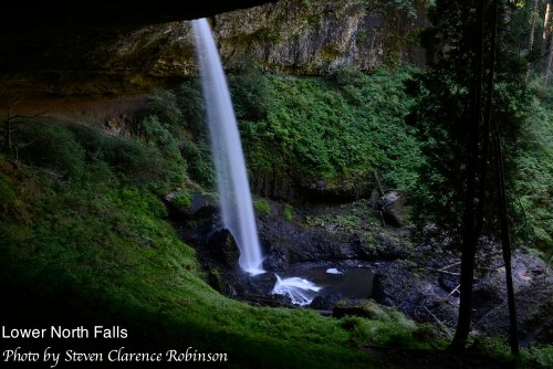 A waterfall cascades into a lush green canyon, surrounded by trees and rocky terrain.