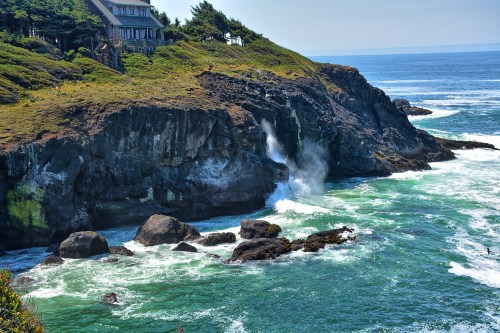 A coastal scene featuring a house on a cliff, waves crashing against rocks, and a clear blue sky.
