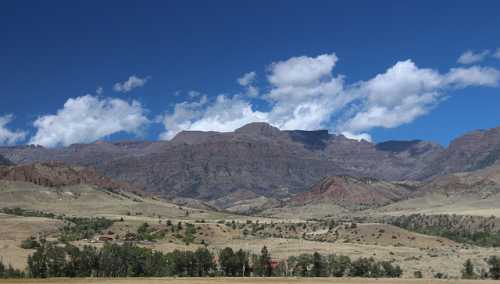 A panoramic view of rugged mountains under a blue sky with scattered clouds, surrounded by dry grassland and trees.