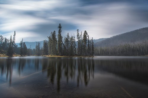 A serene lake surrounded by tall trees, reflecting the cloudy sky and distant mountains in calm waters.
