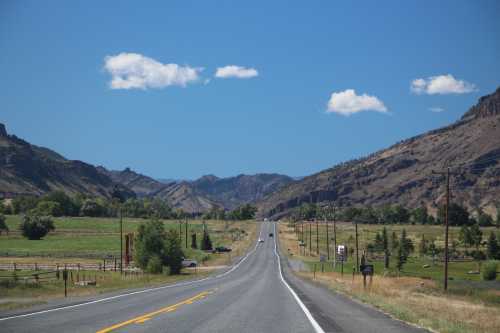 A long, straight road stretches through a scenic landscape of mountains and fields under a clear blue sky.