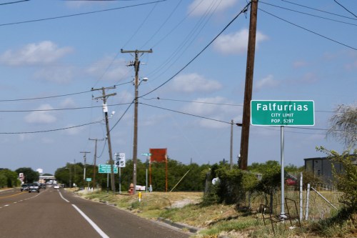Green city limit sign for Falfurrias, Texas, with a population of 5,297, alongside a rural road and power lines.