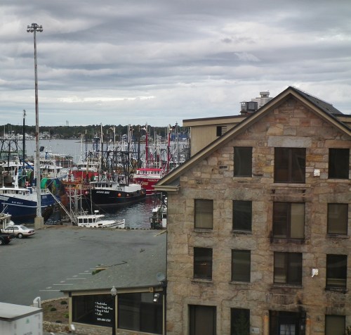 A harbor scene with fishing boats docked near a stone building under a cloudy sky.