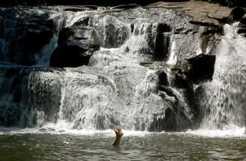 A person with their arms raised is partially submerged in a pool beneath a cascading waterfall.