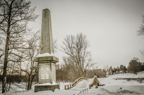 A snowy landscape featuring a tall monument and a wooden bridge, surrounded by bare trees and a serene winter scene.