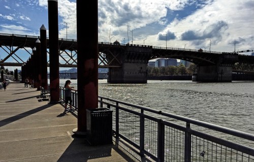 A riverside view featuring a bridge, benches, and a person sitting by the water under a partly cloudy sky.