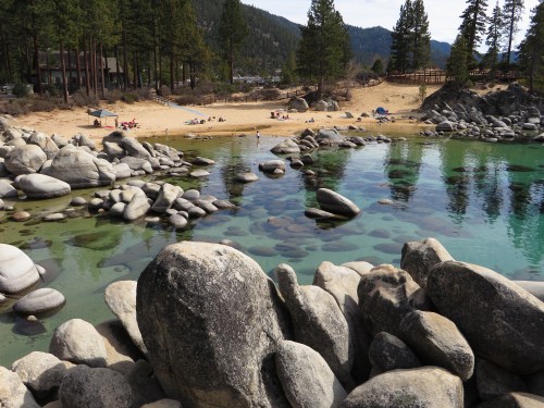 A serene lakeside scene with smooth rocks, clear water, and a sandy beach surrounded by tall trees and mountains.