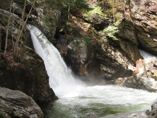 A cascading waterfall flows into a rocky river surrounded by lush greenery and steep cliffs.