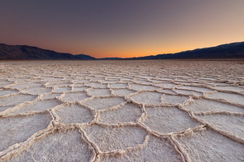 A vast salt flat with hexagonal patterns under a colorful sunset sky and distant mountains.