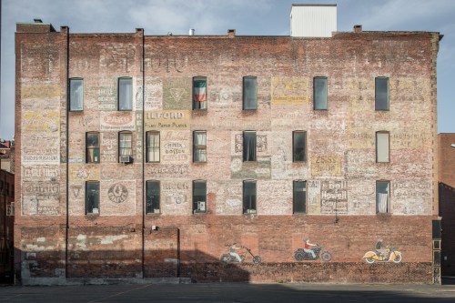 Faded advertisements and murals on a weathered brick building wall, with windows and a shadow in the foreground.