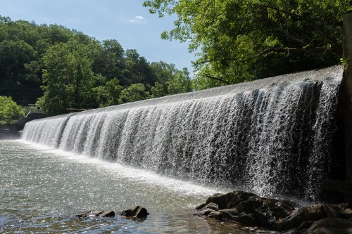 A serene waterfall cascading over a stone dam, surrounded by lush green trees under a clear blue sky.