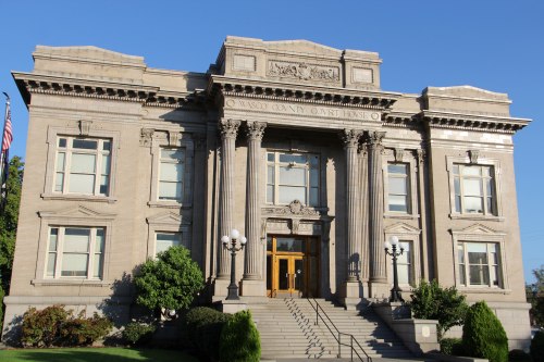 Historic courthouse with grand columns, steps, and decorative details, surrounded by greenery and clear blue sky.