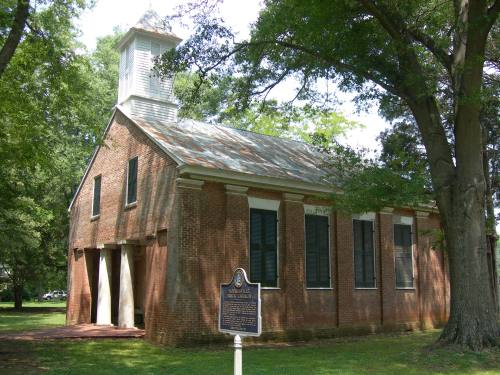 Historic brick building with a bell tower, surrounded by trees and a historical marker in front.