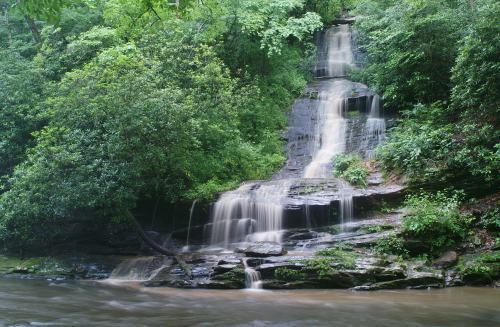 A serene waterfall cascading over rocks, surrounded by lush green foliage and a calm river below.