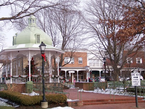 A winter scene featuring a gazebo decorated for the holidays, surrounded by trees and shops in a quaint town square.