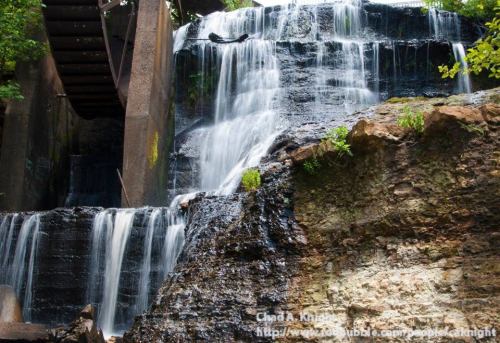 A serene waterfall cascading over rocky terrain, surrounded by lush greenery and a wooden structure above.