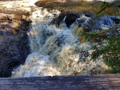 A rushing waterfall cascades over rocks, surrounded by lush greenery and sunlight.