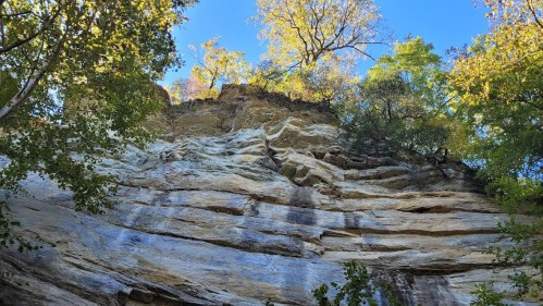 A rocky cliff with layered stone, surrounded by trees and vibrant autumn foliage under a clear blue sky.