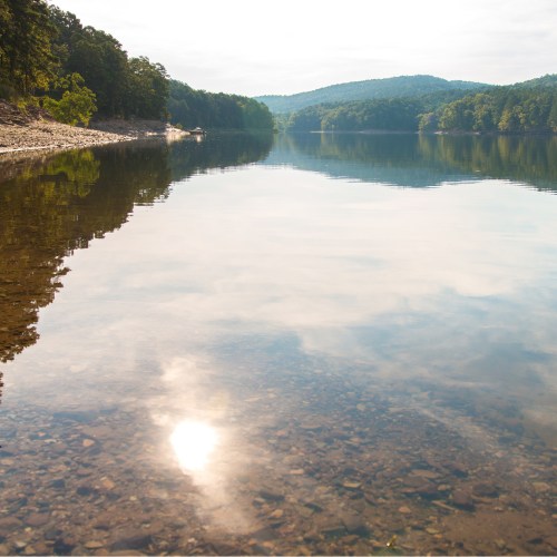 A serene lake reflecting trees and clouds, with a rocky shore and gentle ripples on the water's surface.