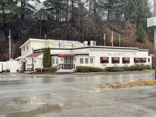 A quaint restaurant with a white exterior, red awnings, and flags, set against a wooded hillside on a rainy day.